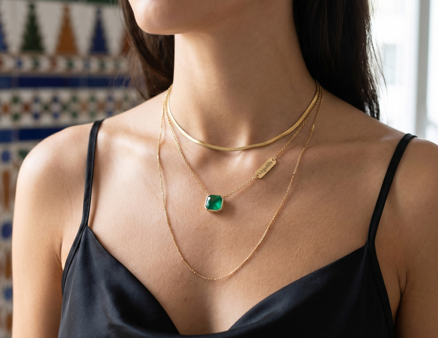 Primer plano de un escote elegante mostrando tres collares de oro superpuestos (tendencia layering) de la marca Virguliilla: una gargantilla lisa, una cadena fina con colgante de esmeralda y una cadena más larga. De fondo, azulejos geométricos andaluces desenfocados.