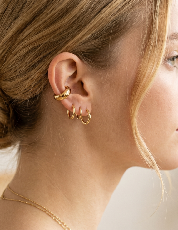 Primer plano de la oreja de una modelo luciendo un ear cuff liso de oro de la marca Virguliilla ajustado en el cartílago, combinado con aros pequeños en el lóbulo.
