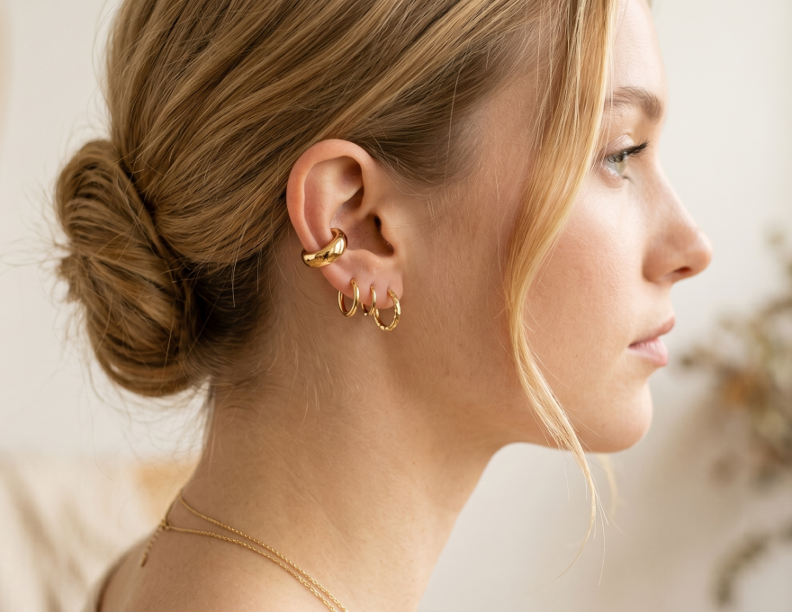 Primer plano de la oreja de una modelo luciendo un ear cuff liso de oro de la marca Virguliilla ajustado en el cartílago, combinado con aros pequeños en el lóbulo.