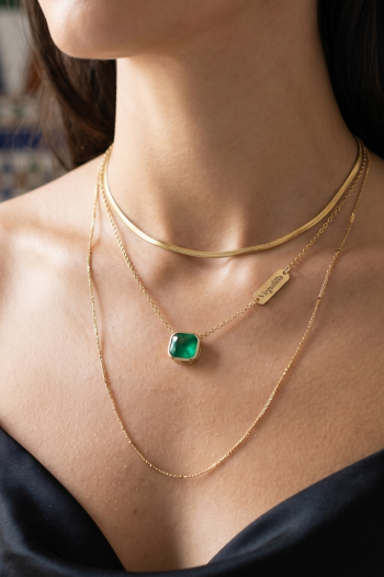 Primer plano de un escote elegante mostrando tres collares de oro superpuestos (tendencia layering) de la marca Virguliilla: una gargantilla lisa, una cadena fina con colgante de esmeralda y una cadena más larga. De fondo, azulejos geométricos andaluces desenfocados.